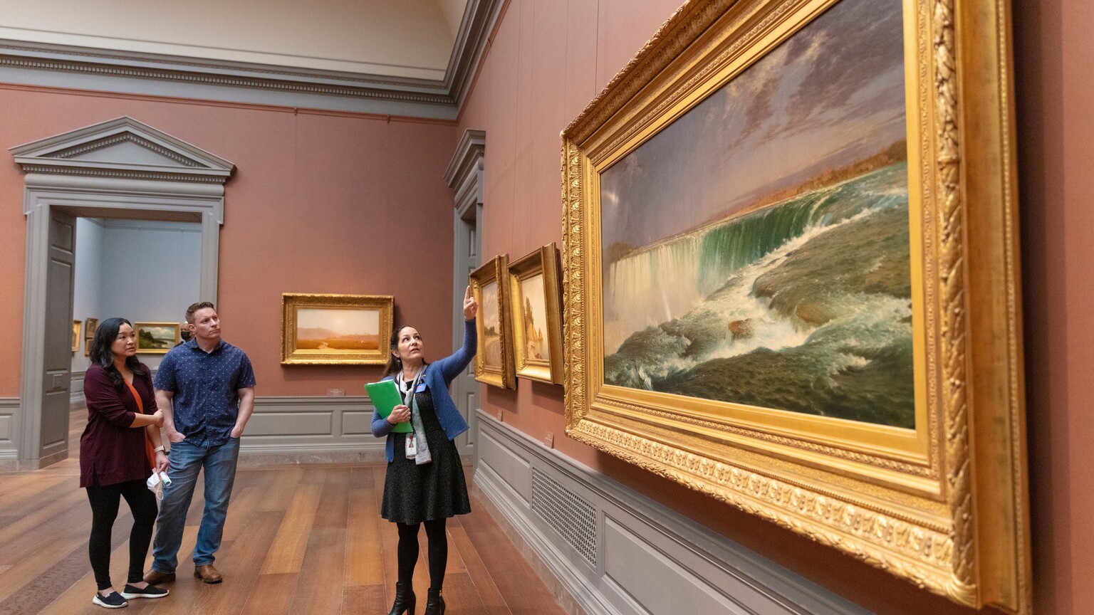 A woman showing and man and a woman a painting in the National Gallery of Art.