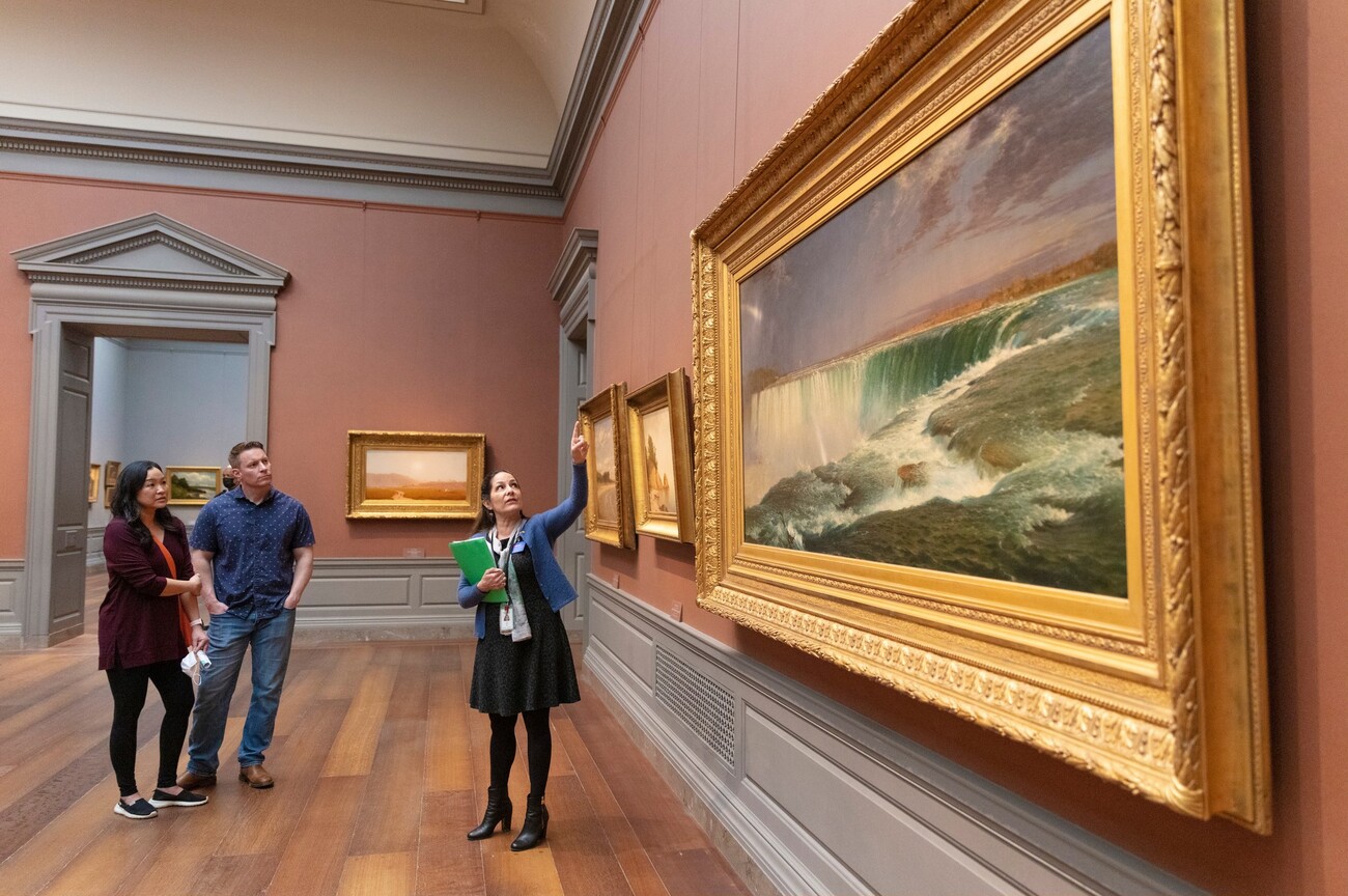 A woman showing and man and a woman a painting in the National Gallery of Art.