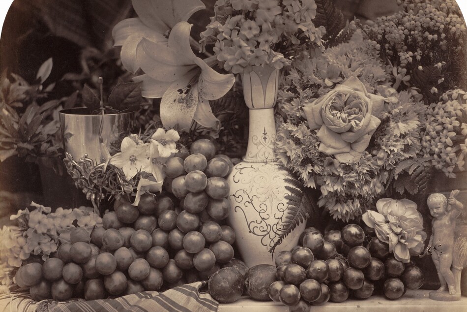 Grapes and flowers arranged on a tabletop in front of a dark background fill this horizontal photograph. The image is monochromatic like a black and white photograph but is printed in warm tones of golden and dark browns. The tabletop runs parallel and close to the bottom edge of the composition. It seems to be made of marble and a striped cloth folds over the front edge of the table to our left. Dark grapes are bunched around a vase at the middle of the composition. The vase's tall oval body and long, tapering neck are painted with decorative scrolls and curlicues. A few pieces of small round fruit, perhaps plums, rest on the table in front of the vase. A gleaming goblet to the left and a small figurine of a young boy with a sheaf of wheat to our right are almost lost in the profusion of roses, lilies, hyacinth, and other flowers that fill the space around and behind the objects. The top corners of the photograph are curved, creating a shallow arch.