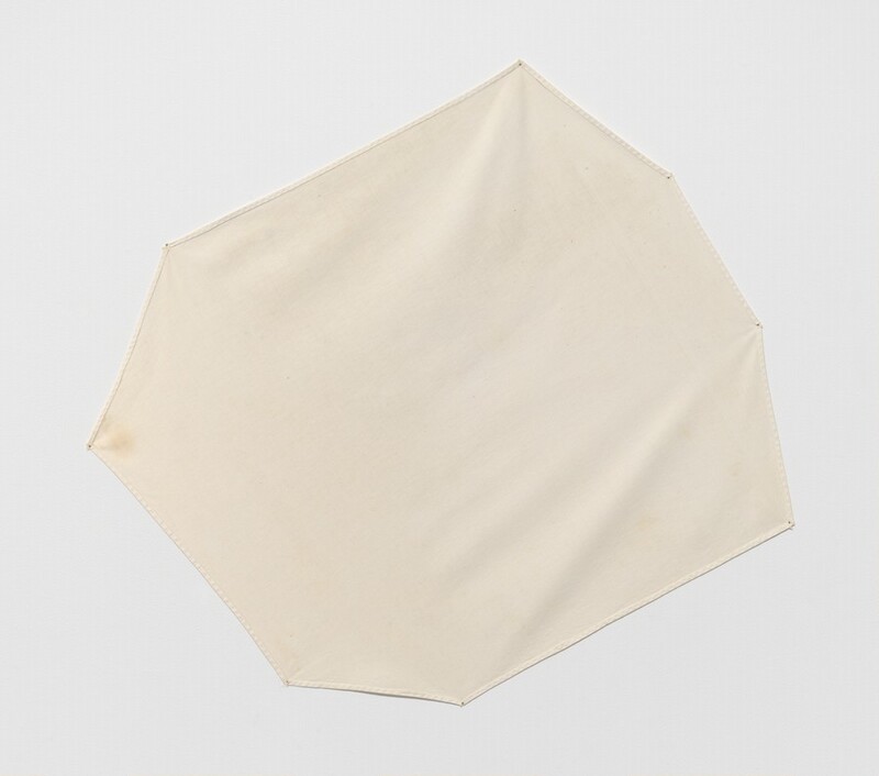 An eight-sided, parchment-white cloth is stretched out and pinned to a wall in this square photograph. The sides are different lengths, and the work is hung angled down to our left. The fabric is puckered in some places, so it casts faint shadows across its surface. There is a light brown stain at the corner near the middle left.