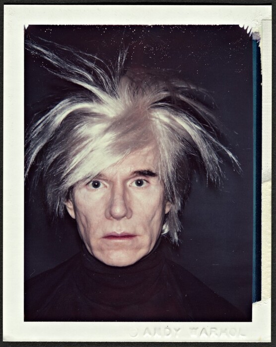 This photograph shows a person's face and upper shoulders. The person is facing forward, looking directly at us. Their hair is silver-grey and wild, with long strands pointing in various directions. The individual's skin is slightly wrinkled, and they have light-colored skin, pronounced cheekbones, deep-set eyes, and thin lips in a neutral expression. They are wearing a black turtleneck garment that blends in with the dark background. The photograph is surrounded by an off-white border.