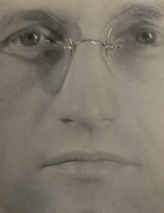The image depicts a close-up view of a person's face, emphasizing the eyes, nose, and mouth, with no other body parts in view. The face fills the frame, with the eyes looking at the camera. The person wears thin-rimmed round glasses, and there are signs of aging around the eyes. The mouth is closed with slightly pursed lips. The image is in black and white, and the background is blurred, focusing solely on the facial features.