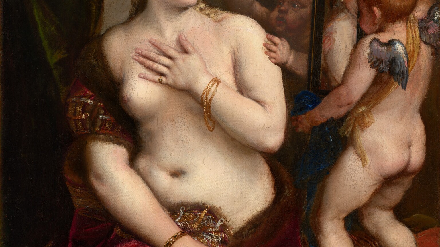 A partially nude woman with pale, peachy skin sits to our left looking into a mirror held up by a nearly nude, winged child to our right in this vertical painting. The woman’s body is angled to our left but she looks across her body to our right. She holds her left hand to her chest, and her right hand grips the fur-lined edge of the scarlet-red, velvet fabric that drapes over that upper arm and around her hips. Her blond hair is coiled up in rows of pearls, and she has dark eyes, a straight nose, and her pale pink lips turn up in a subtle smile. A teardrop pearl earring hangs from the ear we can see, and she wears rings and gold bracelets. The child-like figure to the right stands on a gold-striped cushion facing away from us as he holds up the rectangular, black-framed mirror. He has small silver wings and a sash of golden yellow hangs from one shoulder around the opposite hip. A second child reaches from behind the mirror to hold up a ring of laurel leaves. A forest-green curtain is gathered in the upper left corner and the beige wall beyond falls into shadow to our right.