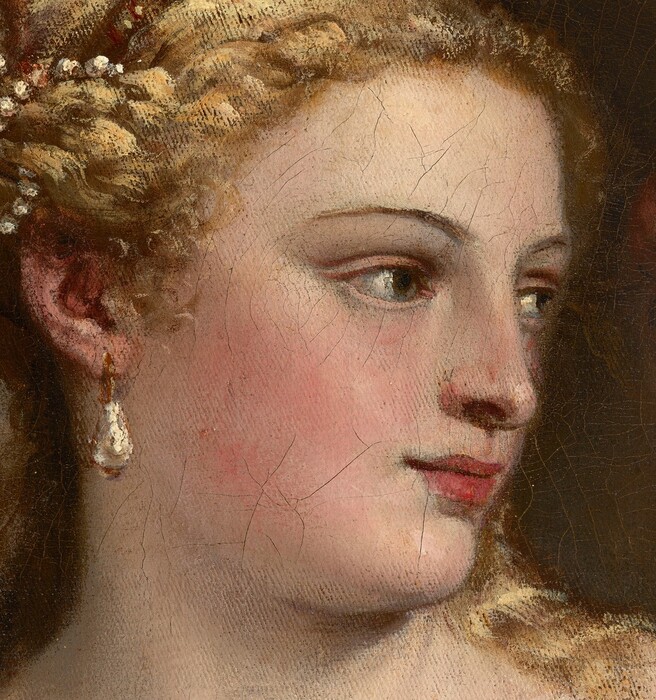 A partially nude woman with pale, peachy skin sits to our left looking into a mirror held up by a nearly nude, winged child to our right in this vertical painting. The woman’s body is angled to our left but she looks across her body to our right. She holds her left hand to her chest, and her right hand grips the fur-lined edge of the scarlet-red, velvet fabric that drapes over that upper arm and around her hips. Her blond hair is coiled up in rows of pearls, and she has dark eyes, a straight nose, and her pale pink lips turn up in a subtle smile. A teardrop pearl earring hangs from the ear we can see, and she wears rings and gold bracelets. The child-like figure to the right stands on a gold-striped cushion facing away from us as he holds up the rectangular, black-framed mirror. He has small silver wings and a sash of golden yellow hangs from one shoulder around the opposite hip. A second child reaches from behind the mirror to hold up a ring of laurel leaves. A forest-green curtain is gathered in the upper left corner and the beige wall beyond falls into shadow to our right.