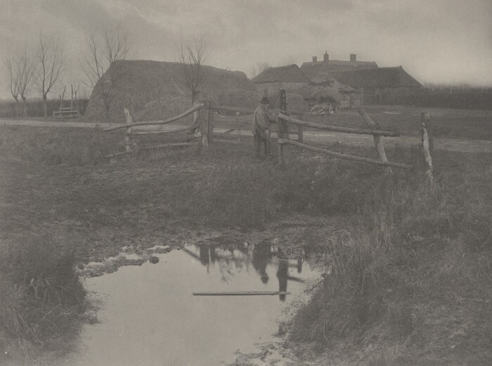 The image shows a low vantage point with a small water puddle reflecting a silhouette of a figure and trees. In the distance, a man stands near a rustic wooden fence separating the close-up area and the field beyond. The horizon is above a line of houses and trees with slanted, thatched roofs, indicating a rural setting. The color palette is monochromatic, dominated by earthy grays and browns suggesting overcast or late afternoon light. The scene lacks vibrant colors, creating a subdued and tranquil atmosphere. The focus is on textures of the wooden fence, grass, and muddied earth.