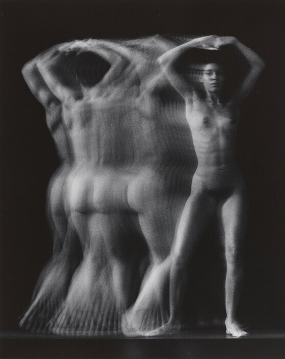 The image shows a full-body depiction of a nude person captured in multiple exposures or a motion blur effect. The person stands with their arms raised above their head, and their legs are slightly apart. The motion blur obscures detailed facial features and gives the hair a soft, indistinct quality. No clothing, jewelry, or objects are visible. The background is a uniform dark shade, which contrasts with the lighter tones of the person's body, emphasizing the movement effect.