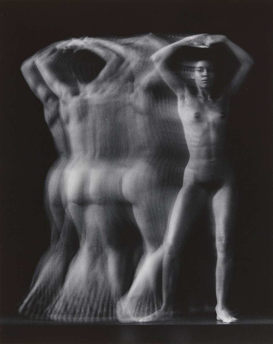 The image shows a full-body depiction of a nude person captured in multiple exposures or a motion blur effect. The person stands with their arms raised above their head, and their legs are slightly apart. The motion blur obscures detailed facial features and gives the hair a soft, indistinct quality. No clothing, jewelry, or objects are visible. The background is a uniform dark shade, which contrasts with the lighter tones of the person's body, emphasizing the movement effect.