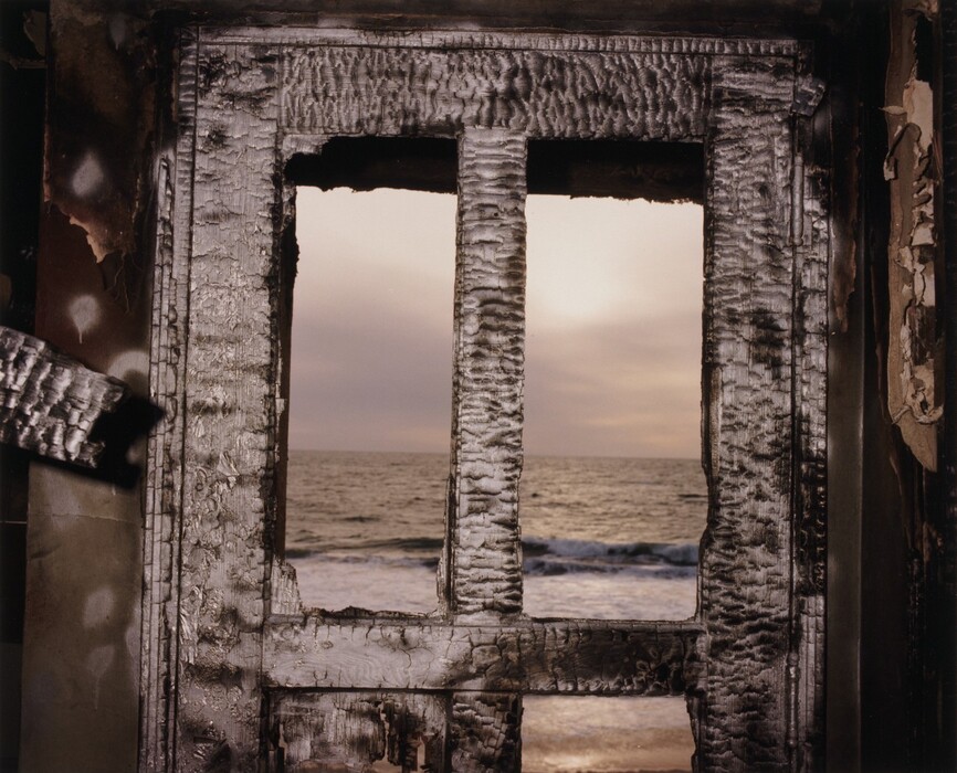 The image shows an interior view through an old, charred window frame with a view of the sea meeting the sky. The color palette includes muted earth tones, soft grays, dusky oranges, yellows, and deep blue-gray. The textures of the burned wood frame the scene. The sandy shore is visible closer to the viewer. The window's deteriorated texture adds a gritty quality.