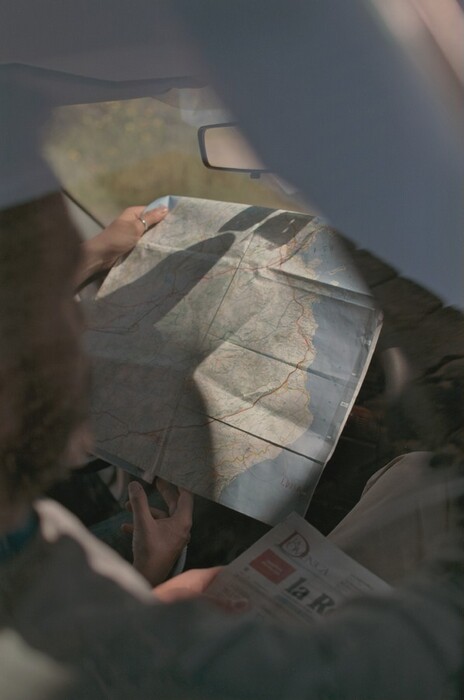 The image shows a map being held and examined inside a vehicle. The composition includes a partial view of the steering wheel, a glimpse of the dashboard, and the reflection of sunlight casting shadows and highlights on the map. The unfolded map displays detailed geographical lines for navigation and is being held near the central area, possibly focusing on a specific location. There is a printed material held beneath the map, like a newspaper. The photo captures a moment of concentration or planning, with the map at the center of the focus.