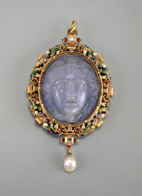 This is an ornate pendant with a carved face. The pendant features a central cameo with a face carved from a semi-transparent, milky blue stone. The face is of a woman, with an open mouth, wide eyes, and curling hair surrounding her face. The carved face is encased in a golden frame with colorful enamel work in blue, green, red, and gold. The pendant has a loop for hanging at the top and a pearl drop hanging from the bottom.