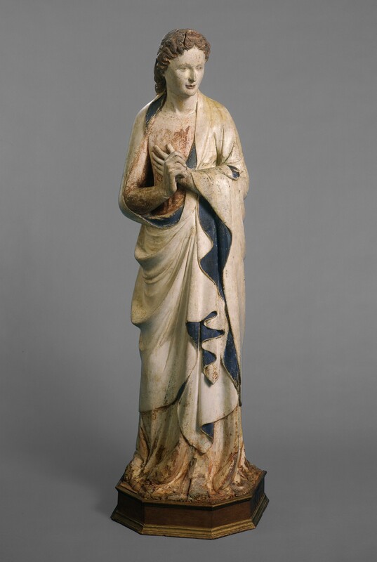 A person stands with hands together at the chest in this freestanding, painted wood sculpture. The person’s body faces us and the head turns and looks slightly down to our right in this photograph. Brown hair curls close to the head and down the back of the neck. The skin is ivory white. The almond-shaped eyes are faintly painted, and he has a straight, delicate nose and thin, slightly smiling pink lips. One hand touches his chest and the other is held in front, perhaps with thumbs touching, palm facing our right. White robes lined with indigo blue fall in cascading folds down his body over a rose-pink tunic, and both pool on the octagonal wood base. There are paint losses and small areas of damage throughout but is most noticeable at the bottom. The wood is cracked in the person’s hair.