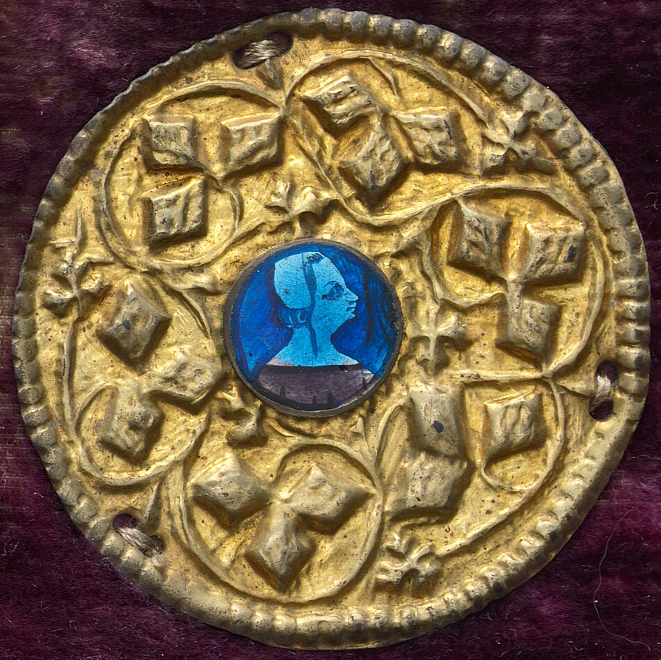 This is a photograph of a circular metallic object. The object features a golden circular medallion with a textured pattern of raised geometric and floral motifs. In the center, there is a small, circular inset with a stylized profile in shades of blue and turquoise against a deep blue background. The object's design suggests a blend of historical and artistic elements. The gold sections have an embossed look reminiscent of decorative craftsmanship, hinting at a ceremonial or symbolic purpose. The medallion's edge is finely scalloped and the background of the photograph is a deep, velvety shade of maroon.