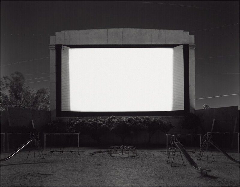 A movie screen is bright white against a dark gray sky, trees, and a playground in this horizontal, black and white photograph. The screen takes up about a quarter of the overall composition, just above the center of the picture. It is raised off the ground, perhaps on a wall or stage that we cannot see. Light from the screen brushes the tops of trees or shrubs growing near the bottom edge. Three sliding boards, several swing sets, and a merry-go-round fade in and out of shadow around the screen’s glow. Power lines crisscross the dark, clear sky behind the screen.