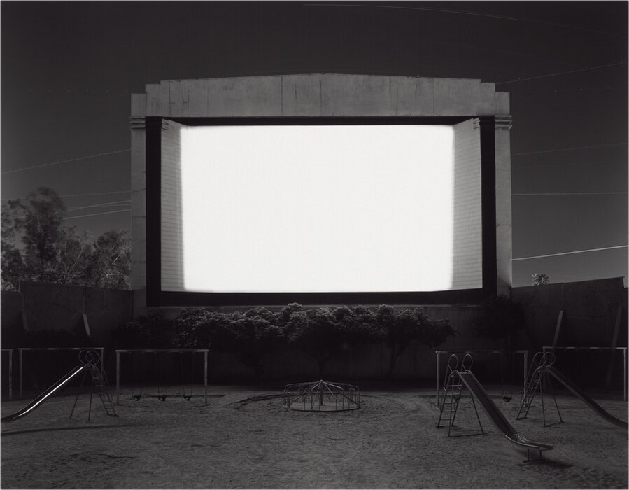 A movie screen is bright white against a dark gray sky, trees, and a playground in this horizontal, black and white photograph. The screen takes up about a quarter of the overall composition, just above the center of the picture. It is raised off the ground, perhaps on a wall or stage that we cannot see. Light from the screen brushes the tops of trees or shrubs growing near the bottom edge. Three sliding boards, several swing sets, and a merry-go-round fade in and out of shadow around the screen’s glow. Power lines crisscross the dark, clear sky behind the screen.