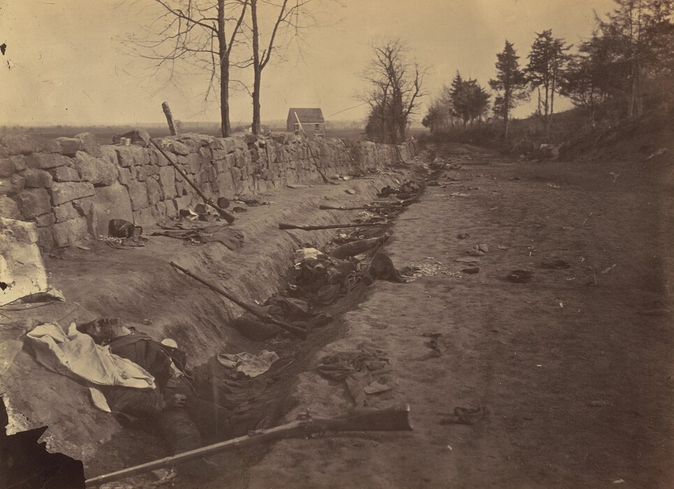 This is a photograph of a battlefield trench lined with objects. The image captures a scene along a dirt trench, bordered by a stone wall on one side, and marked by scattered items like rifles, hats, and pieces of cloth. The stone wall extends into the distance alongside a path that fades into the horizon amid barren trees. The photograph is taken in a sepia tone.