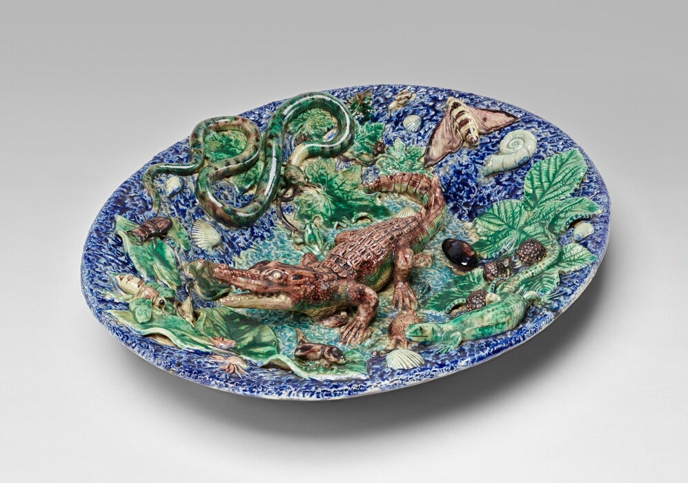 The sculpture is a ceramic dish featuring raised figures from the animal kingdom. At the center is a brown crocodile. Other figures include a coiled snake on the left and a moth on the right. Smaller figures like snails, shells, and leaves are scattered around the dish. The dish is predominantly blue with various colored figures that contrast against the blue background. The piece appears three-dimensional with vividly carved and colored creatures.