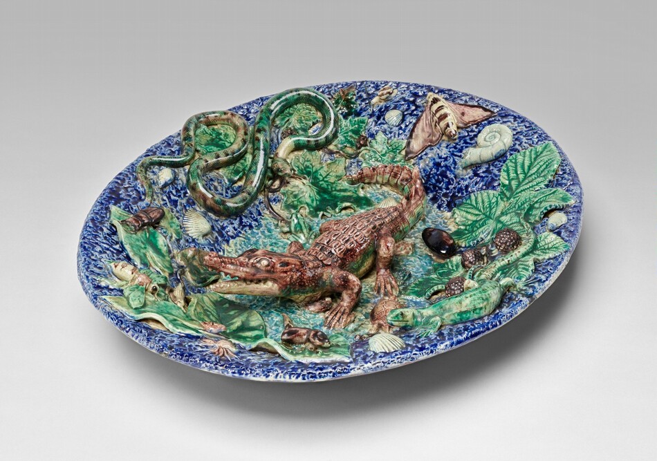 The sculpture is a ceramic dish featuring raised figures from the animal kingdom. At the center is a brown crocodile. Other figures include a coiled snake on the left and a moth on the right. Smaller figures like snails, shells, and leaves are scattered around the dish. The dish is predominantly blue with various colored figures that contrast against the blue background. The piece appears three-dimensional with vividly carved and colored creatures.