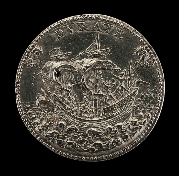 The sculpture depicts a detailed, round antique ship sailing on turbulent waters with the word "DYRATE" inscribed above it. The object appears to be made of a shiny, silver-colored metallic material resembling a coin or medallion, featuring an intricately carved ship with sails, rigging, and waves, all surrounded by a beaded edge. The ship is central to the piece, showcasing careful craftsmanship in its three-dimensional appearance against the flat background.