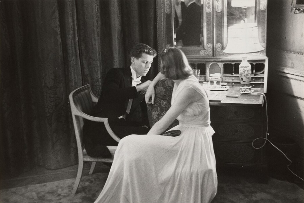 This grayscale photograph shows a man and a woman in an indoor setting. The man is seated, wearing a dark suit with a white shirt. He is leaning forward, reaching towards the woman. She is sitting slightly in front of him in a patterned chair, with one arm resting on the back of the chair and her head turned away from us to look at the man. The woman wears a flowing, short-sleeved white dress, and her hair is styled in a bob. Both people have pale skin. They are sitting in front of a dressing table or vanity, which holds a lamp with a decorative base and other indistinguishable objects. Behind the vanity, there is a large curtain on the left and part of a wall with moldings on the right.