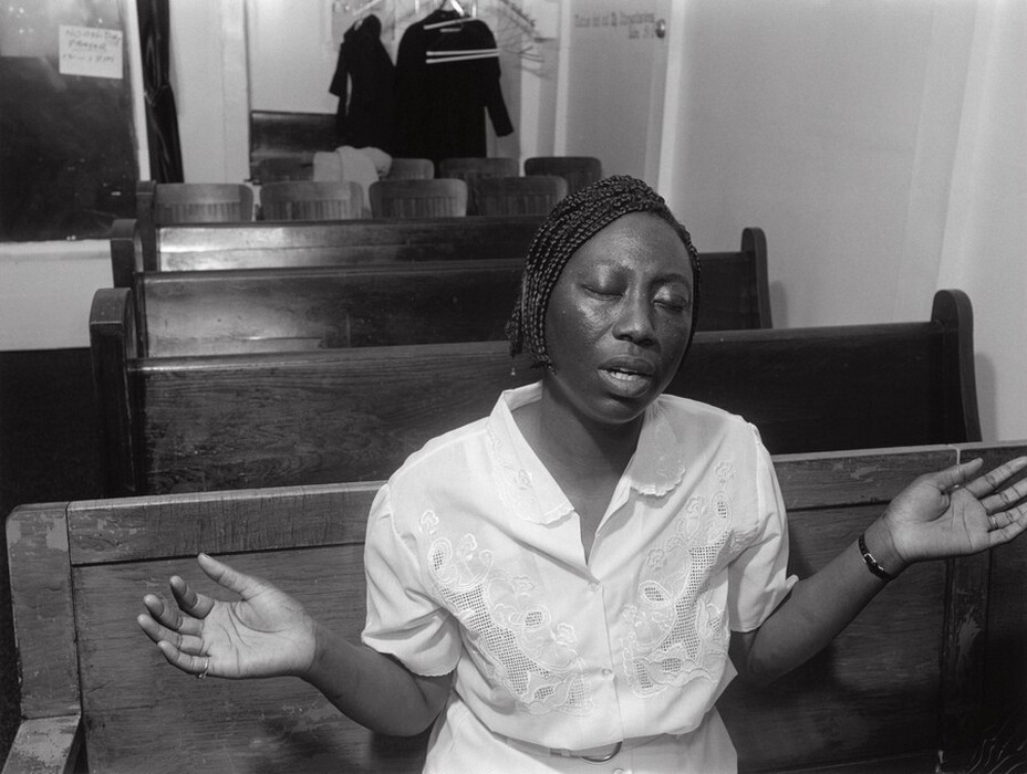 The image shows a woman seated on a wooden pew. Her body is facing forward with her arms slightly raised and hands open. She has braided hair, closed eyes, and a partially open mouth. She is wearing a white shirt with embroidery, a wristwatch on her left wrist, and appears to be wearing a ring on her right hand. The background includes empty wooden pews, a coat rack with garments, and a door with text.