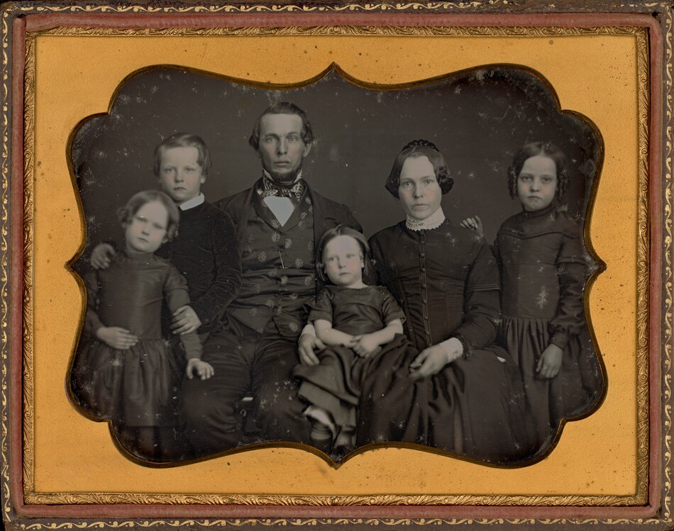 The image shows a vintage portrait of a family consisting of six individuals, all visible from the knees up, sitting and standing closely together. At the center, a man and a woman are seated; the woman holds a young child on her lap. The man has short, neatly combed hair, a prominent forehead, and wears a solemn expression. He is dressed in a formal dark suit with a patterned waistcoat, light cravat, and a high collar shirt underneath. The woman beside him has her hair styled in smooth waves, parted in the center, and wears a serious expression similar to the man. Her attire is a dark dress with a buttoned front and lace detail around the collar. The young child has short hair and is wearing a dress, being supported by the woman. Flanking the seated adults are two girls, standing on either side. Both girls have shoulder-length hair styled in ringlets, wearing long dark dresses similar in style—high neck and long sleeves. Another girl stands on the far left, leaning slightly towards the man, appearing younger than the other two girls, sharing a similar hairstyle. The background of the image is plain, emphasizing the group's presence. The portrait is framed with an ornate, gold-edged mat that complements the subdued tones of the daguerreotype image.