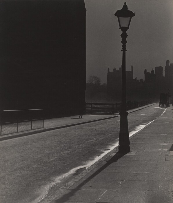 This image shows a street-level view with a curved paved walkway extending into the distance. Various architectural structures create a shadowy background on the right side, contrasted by a tall street lamp in the foreground casting a shadow across the ground. The image features sharp contrasts between light and dark areas, utilizing a monochromatic color palette of black and grey.