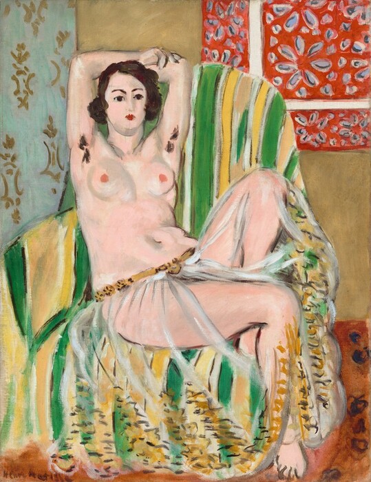 A nearly nude woman with a pale, pink complexion leans back into a striped, upholstered chair with her arms overhead, holding opposite elbows, and one foot tucked under her in this stylized, vertical painting. The scene is painted loosely, making some details indistinct. The woman’s torso faces us but her legs are angled slightly to our right, and she looks off in that direction with dark brown eyes under dark, arched brows. Her features are loosely painted but she seems to have a straight nose and her small, scarlet-red mouth is closed. Her dark brown hair curls down to her ears in a chin-length bob. She wears only a gauzy, transparent cloth, which is gathered low on her hips with a golden-brown belt. Loosely painted, short and zigzag strokes of chocolate brown and harvest gold suggest a wide band of embroidery or decoration around the bottom hem. The woman’s raised arms show two tufts of dark brown hair in her armpits. She has round breasts with dark pink nipples, and a curving belly and hips. Her left knee, farther from us, is bent sharply against the back of the chair so her foot is tucked behind her other thigh. The chair is striped with lime green, white, and lemon yellow. The floor is terracotta orange. A strip of light blue with a gold pattern to the left and a red area with a flower pattern to our right suggest fabrics draped on the sand-brown wall. The artist signed the work in the lower left corner, "Henri Matisse."