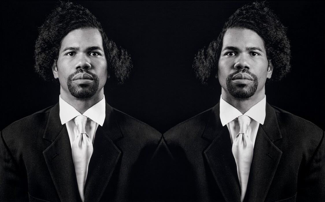 This black-and-white photograph shows a man depicted twice, with only his head and upper torso visible. The depictions appear to be mirrored images. He is facing directly towards the camera with a neutral expression. The man has a dark, closely cropped beard and mustache, medium-toned skin, thick eyebrows, and voluminous, curly hair that is parted dramatically to one side. He is dressed in a formal dark suit jacket over a white dress shirt and white tie. The background is solid black.