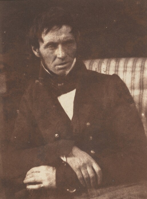 The image shows a man depicted from the waist up. He is seated and leaning slightly forward, with his right hand resting over his left forearm and the fingers of his left hand curled inward. His facial features include a strong jawline, prominent cheekbones, and a focused expression. He has dark hair that appears slightly tousled and unevenly cut. The man is dressed in a dark buttoned coat with a high collar, and it seems that he wears a white shirt underneath, partially visible at the collar. The coat has noticeable buttons that are reflective. There is no visible jewelry, and he is not holding any objects. The background is a simple, abstract surface which includes a fabric with a subtle, likely plaid pattern.