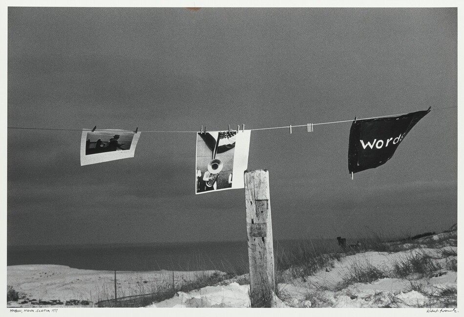Two photographs and a rectangular black banner are pinned to a clothesline above snow-covered sand dunes in this black and white, horizontal photograph. The clothesline spans the width of the composition, and the scene is illuminated by a bright light from the lower right. The photographs on the line and the banner flap in the wind with a dark gray sea and sky looming behind them. The images and banner are secured with clothes pins and spaced across the length of the line. The photograph on the left shows a man wearing a hat and standing at a boat railing looking out at the water. The second photograph is vertically oriented and shows someone playing a tuba, which hides their face, under bunting with stars and stripes. The tuba player is flanked by people on either side who are cropped so only one of their arms is shown. Five empty clothes pins hang between the center photograph and the black banner on the right, where white lowercase lettering reads, “words.” A weathered wooden post stands between us and the clothesline, just to the right of center in the composition. The photograph is inscribed in the lower left margin and reads, “MABOU, NOVA SCOTIA 1977” and is signed by the artist in the lower right margin, “Robert Frank.”
