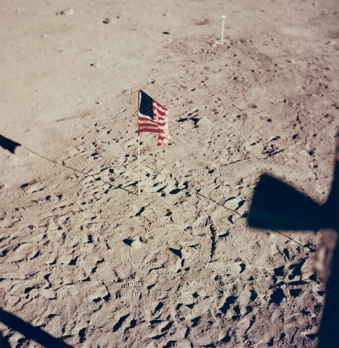 The image is taken from an elevated perspective, showing a rugged, dusty surface. The horizon is not visible, and the focus is on the ground below. In the center is an American flag standing erect, casting a shadow. Around the flag are tire and boot imprints, adding texture. In the lower right, the shadow of a piece of machinery or spacecraft is visible. The color palette is subdued with the flag's colors standing out.