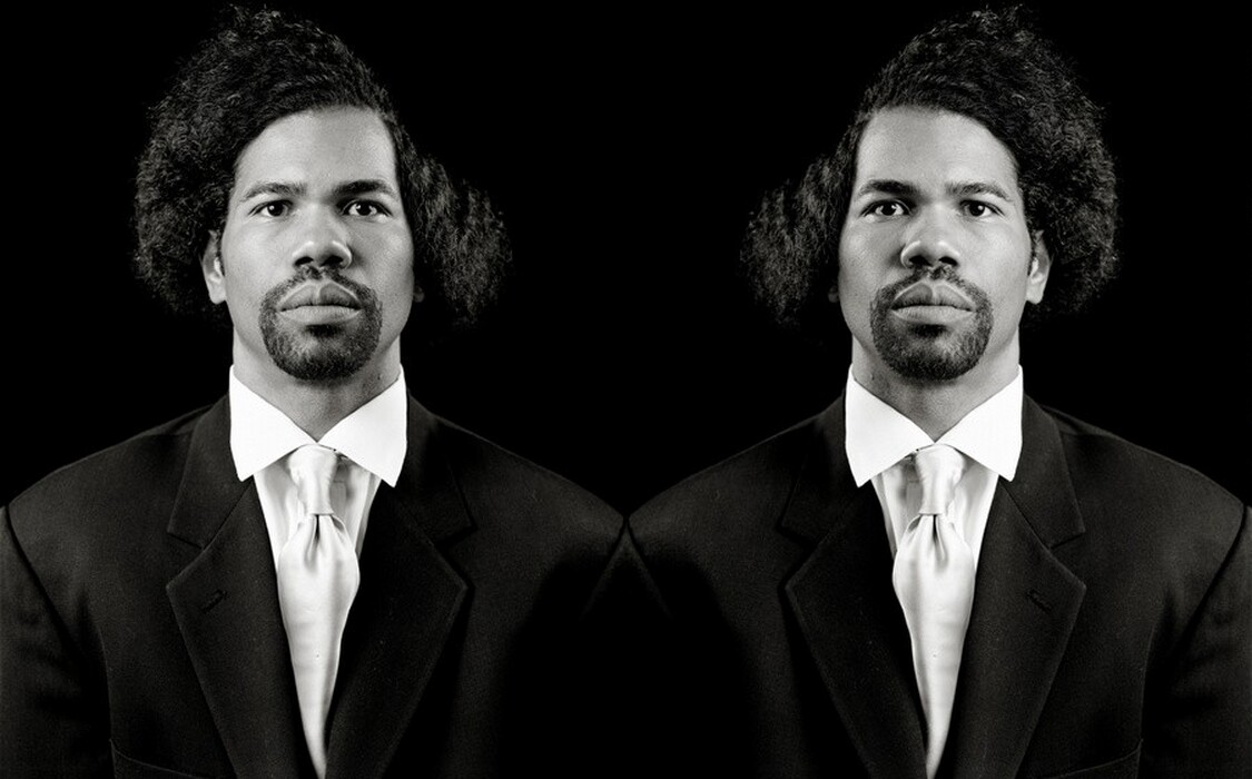 This black-and-white photograph shows a man depicted twice, with only his head and upper torso visible. The depictions appear to be mirrored images. He is facing directly towards the camera with a neutral expression. The man has a dark, closely cropped beard and mustache, medium-toned skin, thick eyebrows, and voluminous, curly hair that is parted dramatically to one side. He is dressed in a formal dark suit jacket over a white dress shirt and white tie. The background is solid black.