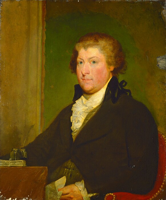 The image is a painted portrait of a man seated in a chair. The man has fair skin, a round face, and curly brown hair. He is wearing a dark coat with a high collar over a white ruffled shirt, with a hint of a patterned vest showing. He is holding a white cloth in his hand on his lap. Beside him, there is a table with papers, an inkstand, and quills. The background is a muted green with texture.