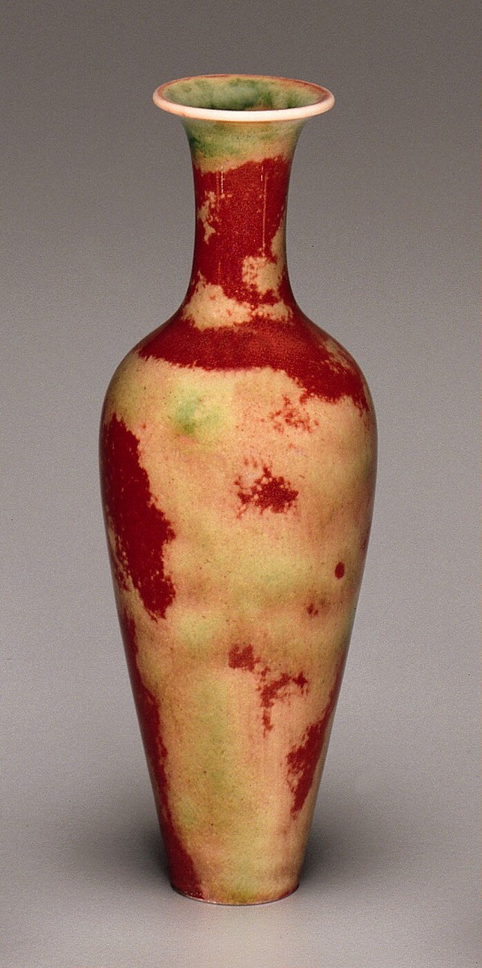 This is a tall ceramic vase with a narrow neck and a flared rim at the top, curving into a rounder body that tapers down towards a narrow bottom. The vase has a mottled mix of deep red, pale yellow, and light green hues. The rim is white, and the interior appears light green.