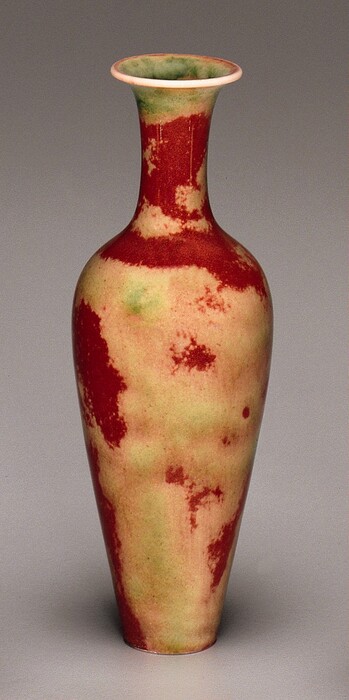 This is a tall ceramic vase with a narrow neck and a flared rim at the top, curving into a rounder body that tapers down towards a narrow bottom. The vase has a mottled mix of deep red, pale yellow, and light green hues. The rim is white, and the interior appears light green.