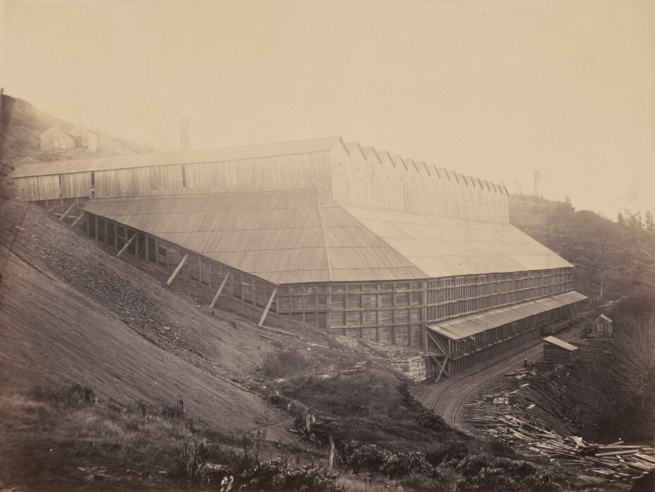 The image shows a large industrial structure on a sloped terrain. The structure is made of wood with vertical beams supporting it. There are smaller buildings in the distance on higher ground to the left, and barren hilltops with sparse trees on the right. The sky appears hazy or overexposed. The color palette is sepia tones.