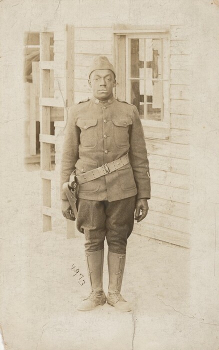 The image shows a man in a military uniform standing upright. He is wearing a jacket with insignia, trousers, and knee-high boots. He also wears a cap and a belt with a holster. The man has a focused expression and closely cropped hair. The background includes a wooden structure with a window and a porch.