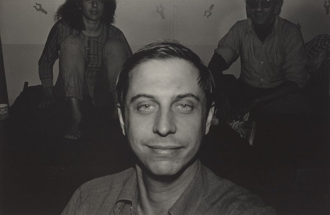 The image shows a man's head and shoulders in the foreground. He has short, light-colored hair, distinct facial features, and is wearing a light-colored collared shirt. In the background, there is a woman with long hair wearing a checkered top and pants, and a man with sunglasses sitting cross-legged. The wall behind them is plain with small, unclear elements or patterns. The lighting highlights the man in the foreground, with the other individuals in shadow.