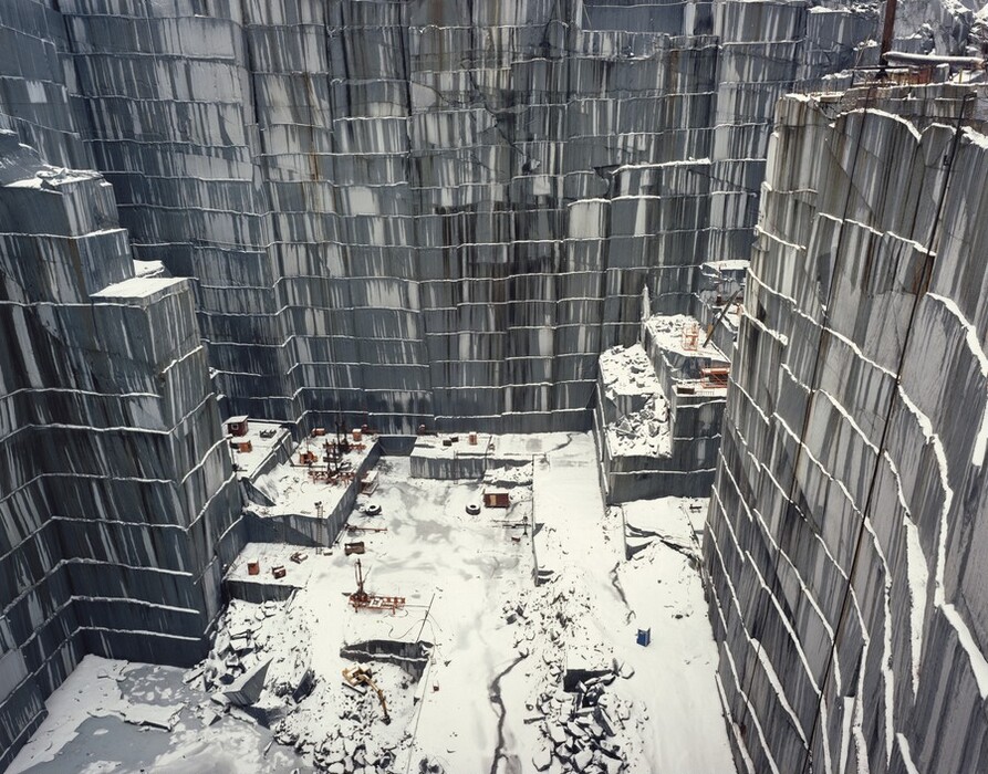 We look down into a straight-sided quarry at mining equipment that appears minuscule in scale in this nearly square color photograph. The walls are streaked with pale and dark gray, and snow rests on narrow ledges between the quarried levels and on the ground. At first glance we might think we’re looking down the sides of skyscrapers onto a city block.