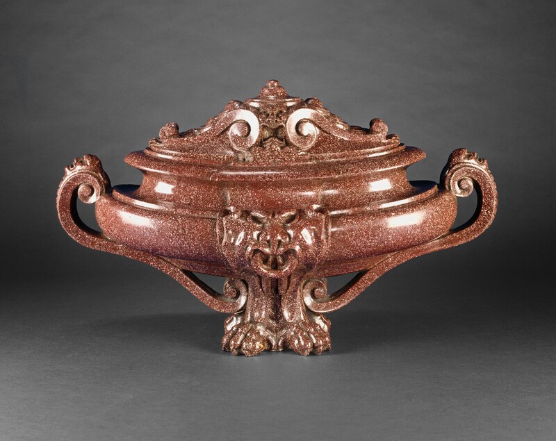 Carved with highly polished, speckled, rust-colored stone, this wide, elongated urn is oval shaped when seen from above. We look onto one longer side in this photograph. The foot of the urn is carved into two lion’s feet, turned slightly outward at an angle from each other. Where the body of the urn meets the feet is an animal-like face with a prominent lower lip and lion-like nose. The lower portion of the urn curves up to create a shallow bowl shape before angling inward into a deeply carved cove above. Two scrolling handles, one on each narrow end of the urn, curve from the bulging bowl of the urn down to the feet. Each handle is decorated with a human face, seen in profile with mouth open, near where the handle meets the body of the urn. The lid is decorated with scrolls and foliation that flank a humanoid face with a gaping mouth, a broad nose, and wide-open eyes. Light glints off the smooth polished surface, and the urn sits against a charcoal-gray background.