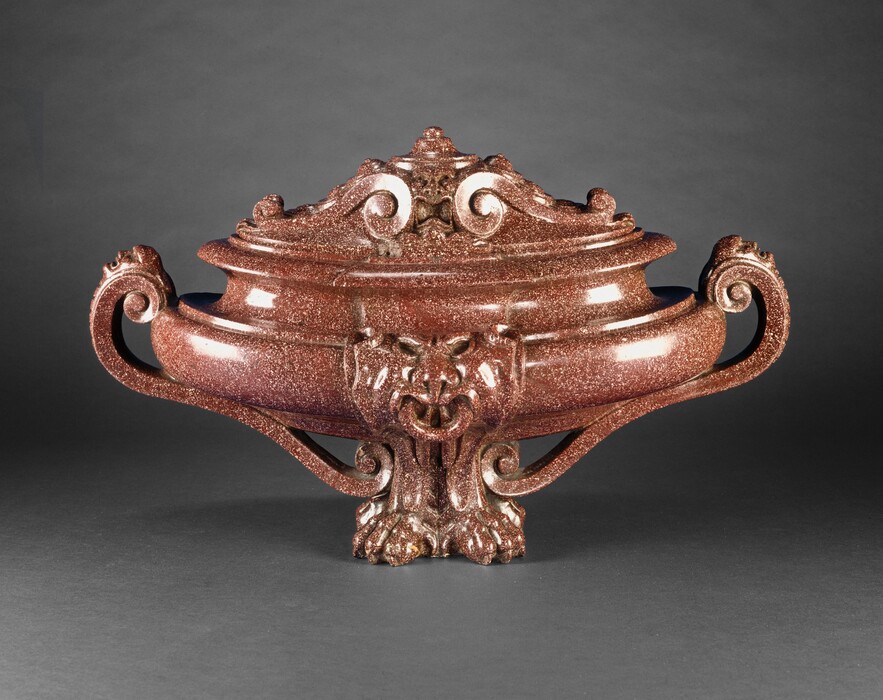Carved with highly polished, speckled, rust-colored stone, this wide, elongated urn is oval shaped when seen from above. We look onto one longer side in this photograph. The foot of the urn is carved into two lion’s feet, turned slightly outward at an angle from each other. Where the body of the urn meets the feet is an animal-like face with a prominent lower lip and lion-like nose. The lower portion of the urn curves up to create a shallow bowl shape before angling inward into a deeply carved cove above. Two scrolling handles, one on each narrow end of the urn, curve from the bulging bowl of the urn down to the feet. Each handle is decorated with a human face, seen in profile with mouth open, near where the handle meets the body of the urn. The lid is decorated with scrolls and foliation that flank a humanoid face with a gaping mouth, a broad nose, and wide-open eyes. Light glints off the smooth polished surface, and the urn sits against a charcoal-gray background.