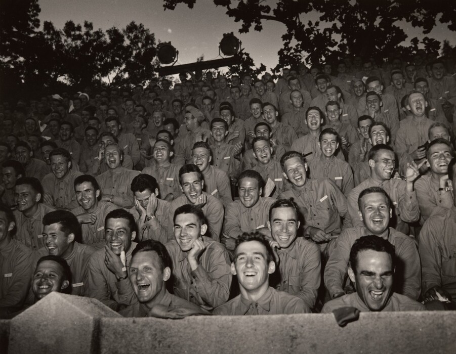 This is a photograph of a large group of individuals seated outdoors, likely watching an entertainment event. The image captures the group laughing and enjoying themselves. The individuals are dressed in military attire and appear to be having a good time. The setting suggests an organized event with overhead lighting indicating nighttime or evening. The photograph is in black and white.
