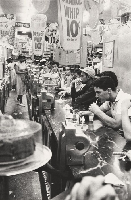 This is a grayscale photograph featuring a diner or soda fountain scene. The photograph shows a long counter stretching away from us with patrons seated on stools, facing the left. Items such as glasses, napkins, and utensils are visible on the counter. Some women stand behind the counter, wearing dresses and headbands. Advertisements for "Orange Whip" for 10 cents are displayed several times in the establishment.
