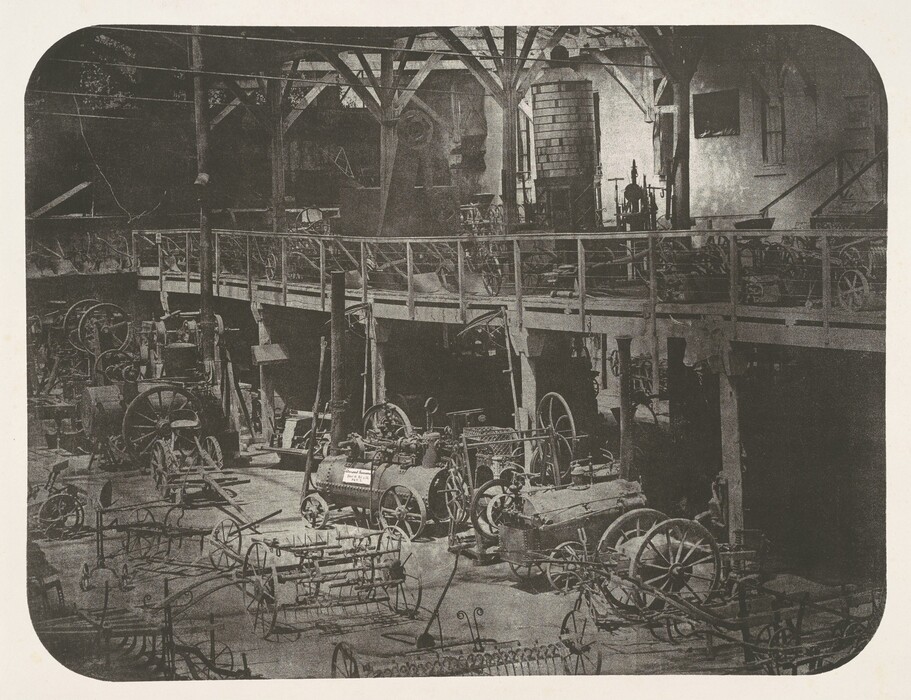 This is a photograph of an indoor space filled with various industrial machinery. The photograph captures a large room with a number of machinery, gears, and equipment. The machinery is arranged across both the floor and an elevated platform supported by wooden columns. The lighting in the photograph suggests it is taken during the daytime, as there is light filtering through unseen windows or openings, illuminating the machinery with shadows. The composition provides a detailed view of the shapes and designs of the devices. Each piece of machinery is distinct, demonstrating different sizes and configurations.