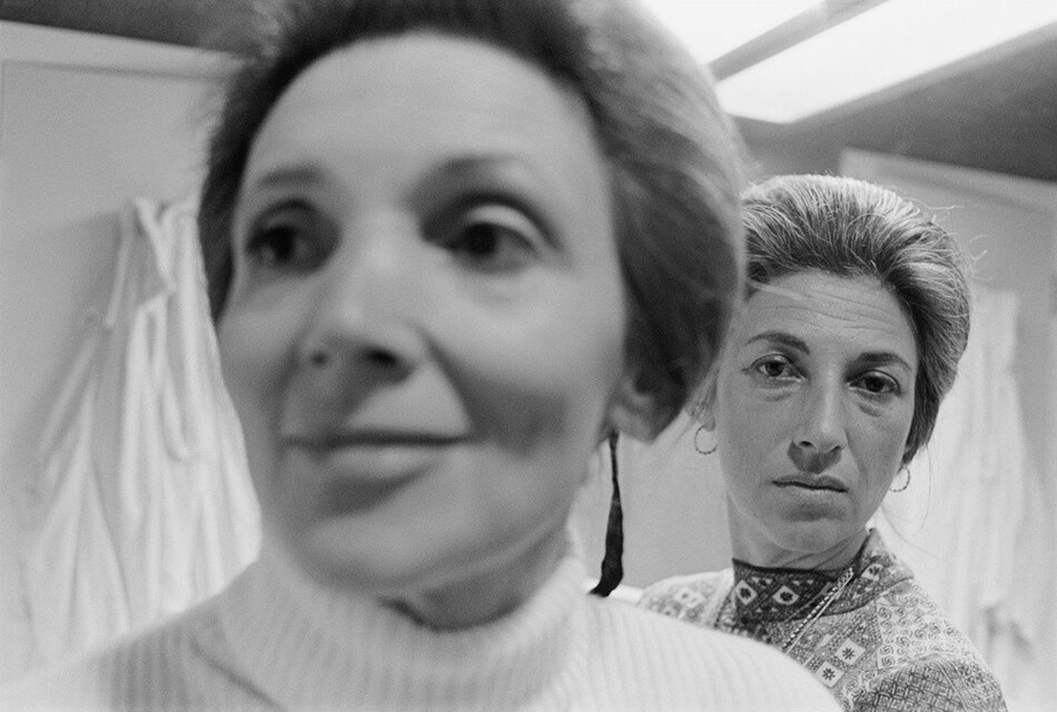 This grayscale photograph shows two women with their upper bodies visible. The woman on the left is closest to us. She is out of focus and is looking slightly to the left, wearing a light-colored, high-necked sweater. The woman on the right is in the background and is in focus, looking directly at the camera. She has her hair pulled back and is wearing a patterned top, hoop earrings, and a necklace. Both women appear to have pale skin and graying hair. The background is indoors, with white garments hanging on the wall. The lighting is overhead and casts shadows.