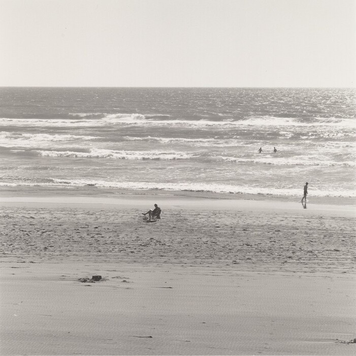 The image displays a seaside view from the sandy shore towards the ocean. The horizon line separates the sky from the sea. The sand shows texture and ripples. A person pushes a stroller, and another figure walks by the water, leaving footprints. Waves break, spreading foam, with two additional figures in the distance. It is captured in monochromatic tones of black, white, and gray, creating a timeless and serene atmosphere.