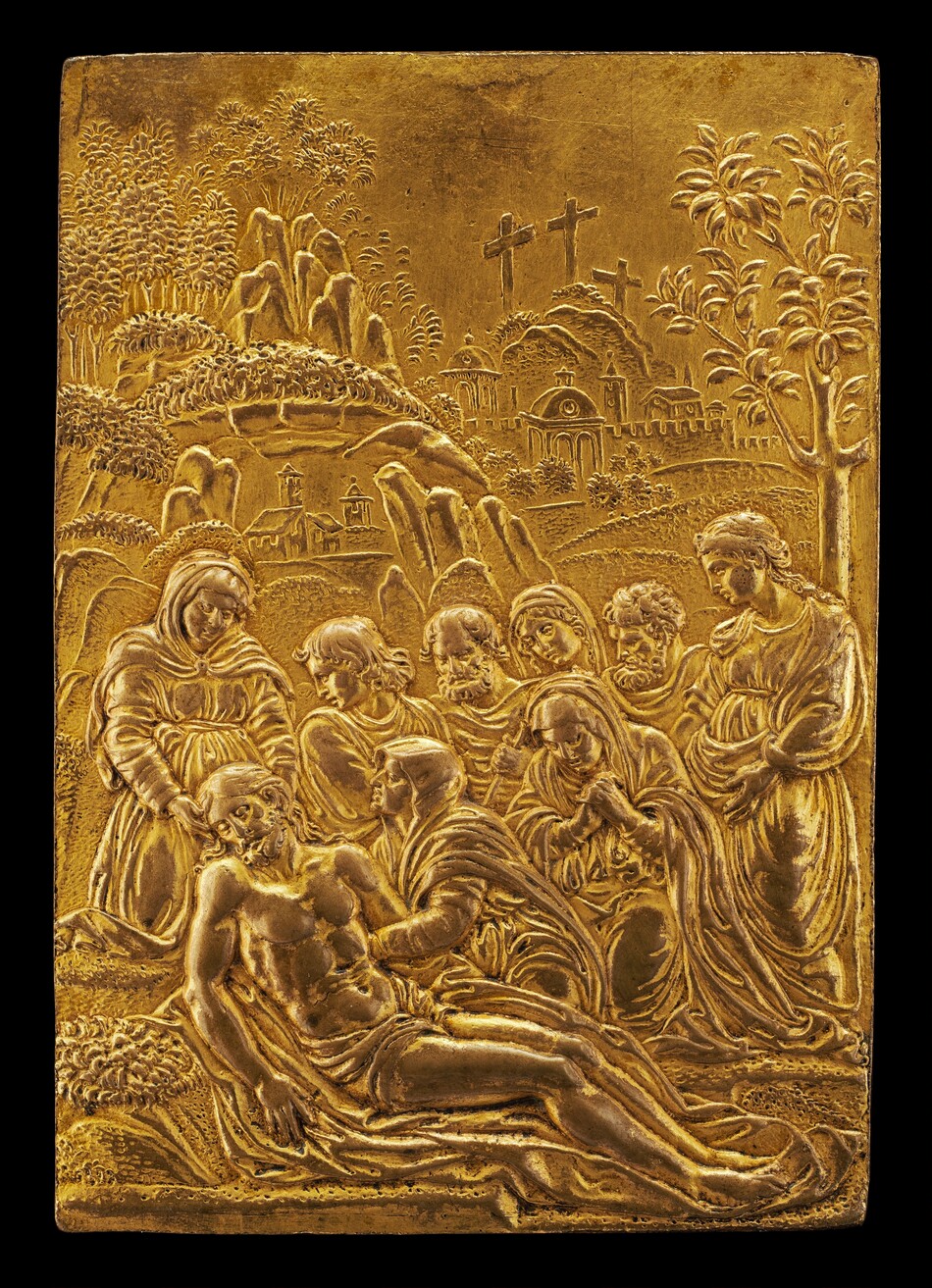 The sculpture shows a group of figures surrounding a central reclining figure, appearing to be a male with defined musculature. The figures are depicted in various poses, some with downcast expressions. All figures wear draped garments. In the background, there are hills, trees, a walled city, and three crosses. The sculpture is made of golden material, giving it a rich appearance. It is crafted as a relief, with figures and landscape elements subtly protruding from the surface.