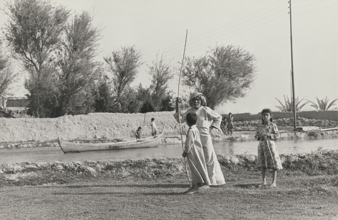 The image shows a scene where an elderly man is talking to a child, while a young girl looks towards the camera. In the background, there is a boat on a body of water. Trees, a mud wall, and a clear sky complete the background. The image is in black and white and includes some modern elements like electrical lines and a pole.