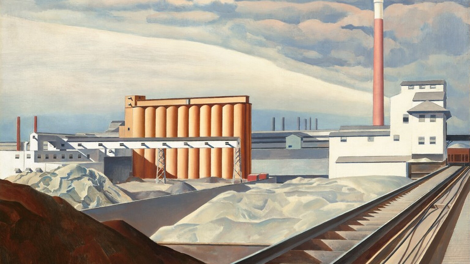 This nearly square painting shows an industrial area with buildings, storage silos, a smokestack, and railroad tracks. A mound of brown dirt or other material is in shadow in the lower left corner of the painting. Next to the mound, railroad tracks extend diagonally from the lower center of the painting into the distance to our right. The tracks end at a white building with staggered gray rooflines to our right in the distance. A tall terracotta-red smokestack rises high beyond the white building, smoke pouring out of its top and blending into the clouds above. Just beyond the mound of dirt, piles of white material, perhaps in unseen bins, line the railroad track to our left and lead back to a row ten interconnected, coral-orange silos. The horizon comes about halfway up the painting, and it is lined with a row of long white and gray warehouses. The artist signed and dated the work with brown paint in the lower right corner: “Sheeler 31.”
