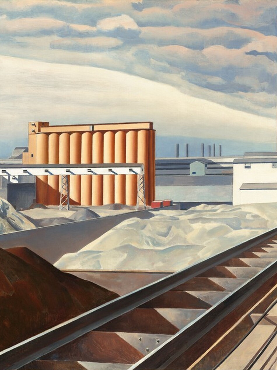 This nearly square painting shows an industrial area with buildings, storage silos, a smokestack, and railroad tracks. A mound of brown dirt or other material is in shadow in the lower left corner of the painting. Next to the mound, railroad tracks extend diagonally from the lower center of the painting into the distance to our right. The tracks end at a white building with staggered gray rooflines to our right in the distance. A tall terracotta-red smokestack rises high beyond the white building, smoke pouring out of its top and blending into the clouds above. Just beyond the mound of dirt, piles of white material, perhaps in unseen bins, line the railroad track to our left and lead back to a row ten interconnected, coral-orange silos. The horizon comes about halfway up the painting, and it is lined with a row of long white and gray warehouses. The artist signed and dated the work with brown paint in the lower right corner: “Sheeler 31.”